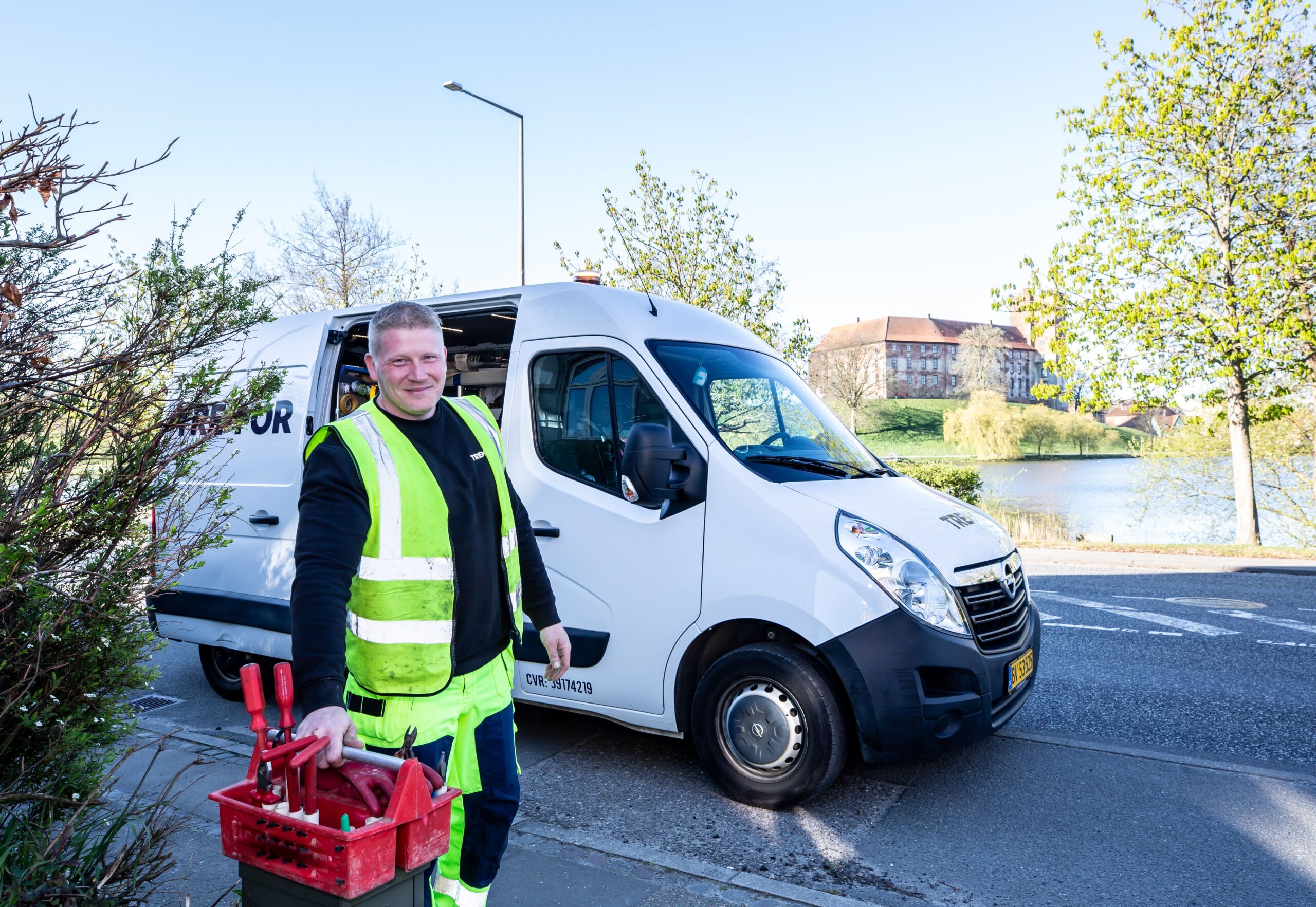 Smiling worker in reflective vest stands by a van holding tools.